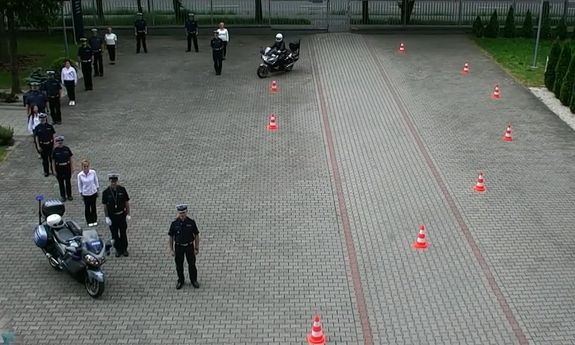 policjanci i pracownicy cywilni stoją po lewej stronie układając kształt połowy serca. Po prawej stronie motocyklista jedzie między pomarańczowymi słupkami, które układają drugą połowę serca