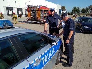 Funkcjonariusze przy radiowozie na tle pojazdów Straży Pożarnej