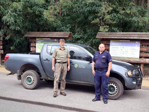 Policjant wspólnie ze strażnikiem Kampinoskiego Parku Narodowego na tle lasu i pojazdu terenowego Kampinoskiego Parku Narodowego