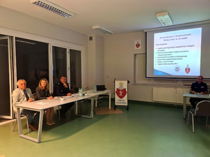Przedstawiciele Policji u Urzędu Gminy w Kampinosie podczas debaty społecznej siedzą za stołem , w tle prezentacja multimedialna na temat bezpieczeństwa w gminie