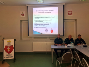 Przedstawiciele Policji podczas debaty społecznej siedzą za stołem, w tle prezentacja multimedialna na temat bezpieczeństwa w gminie