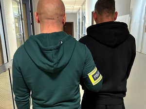 Nieumundurowany policjant z opaską na ręce z napisem "Policja" doprowadza zatrzymanego mężczyznę do jednostki